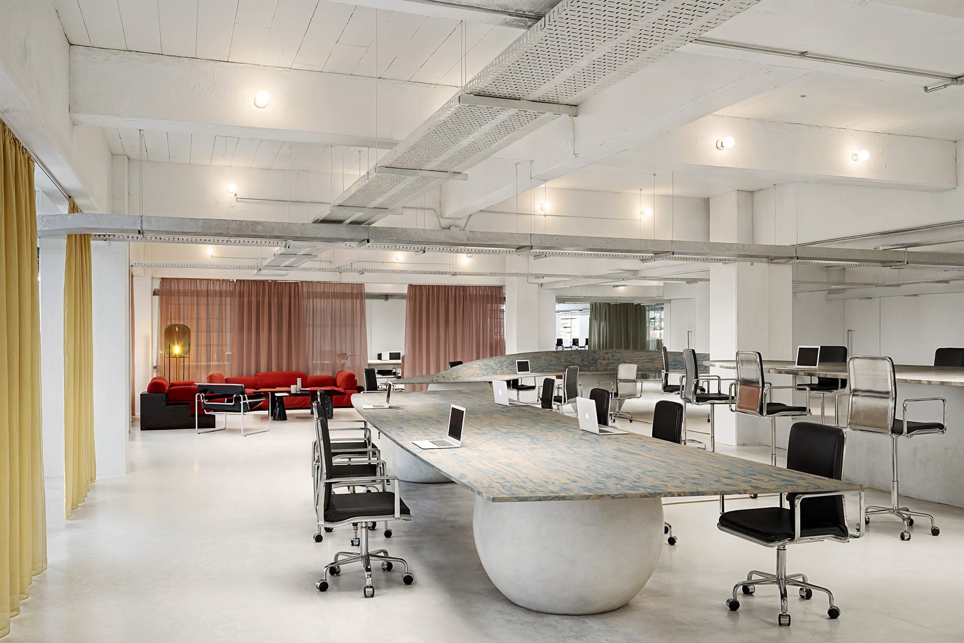 Spacious modern office with long tables, chairs, and red couches in the background.