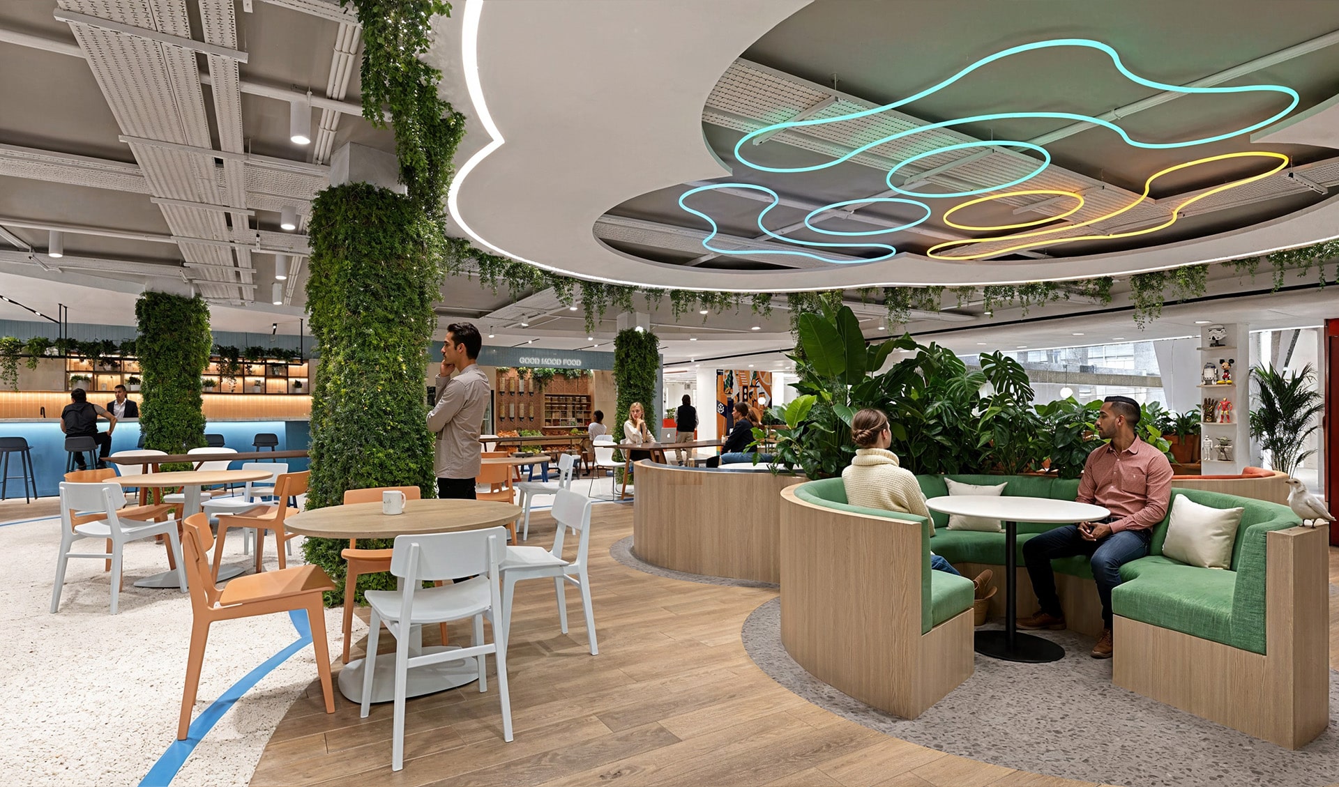 Modern cafe with green decor, round seating, and neon ceiling lights. People are seated and chatting.